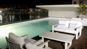 Área de piscina en la azotea con sillas de descanso blancas y una mesa, con vista a una masa de agua de noche.
