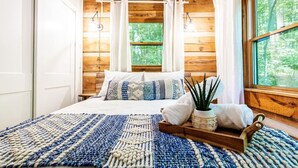 Ein gemütliches Schlafzimmer mit einem Bett, einem blau-weißen Teppich, einem hölzernen Kopfteil und einem Fenster mit Blick auf Bäume.