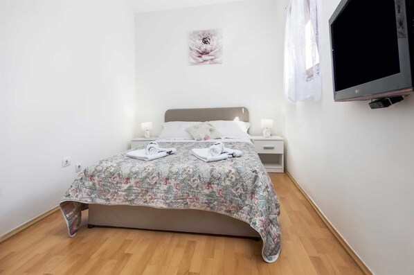 Un lit bien fait, avec une literie à motifs floraux, un téléviseur fixé au mur et une simple table de chevet avec une lampe.