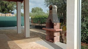 Un espace barbecue extérieur doté d’une cheminée en pierre, entouré de verdure et comprenant un coin salon abrité.