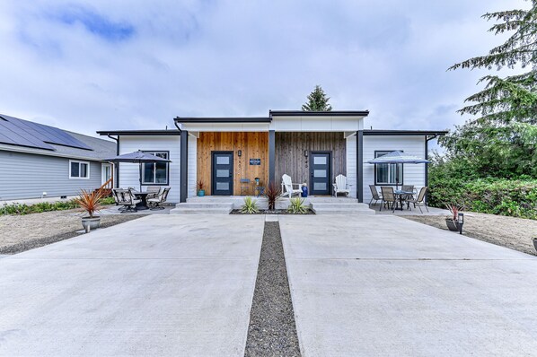 Une maison moderne à deux étages, dotée d’une porte d’entrée en bois, d’un patio couvert avec des meubles d’extérieur et d’un chemin de gravier menant à l’entrée.