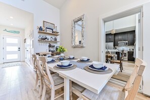 Un coin repas avec une table blanche dressée pour six personnes, des chaises en bois et une cuisine visible à travers une porte ouverte.