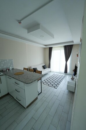 A modern kitchen with white cabinets and granite countertops, a dining area with a sofa and a rug, and a large window with curtains. - İndag Apart Aile Oteli  (Seferihisar)
