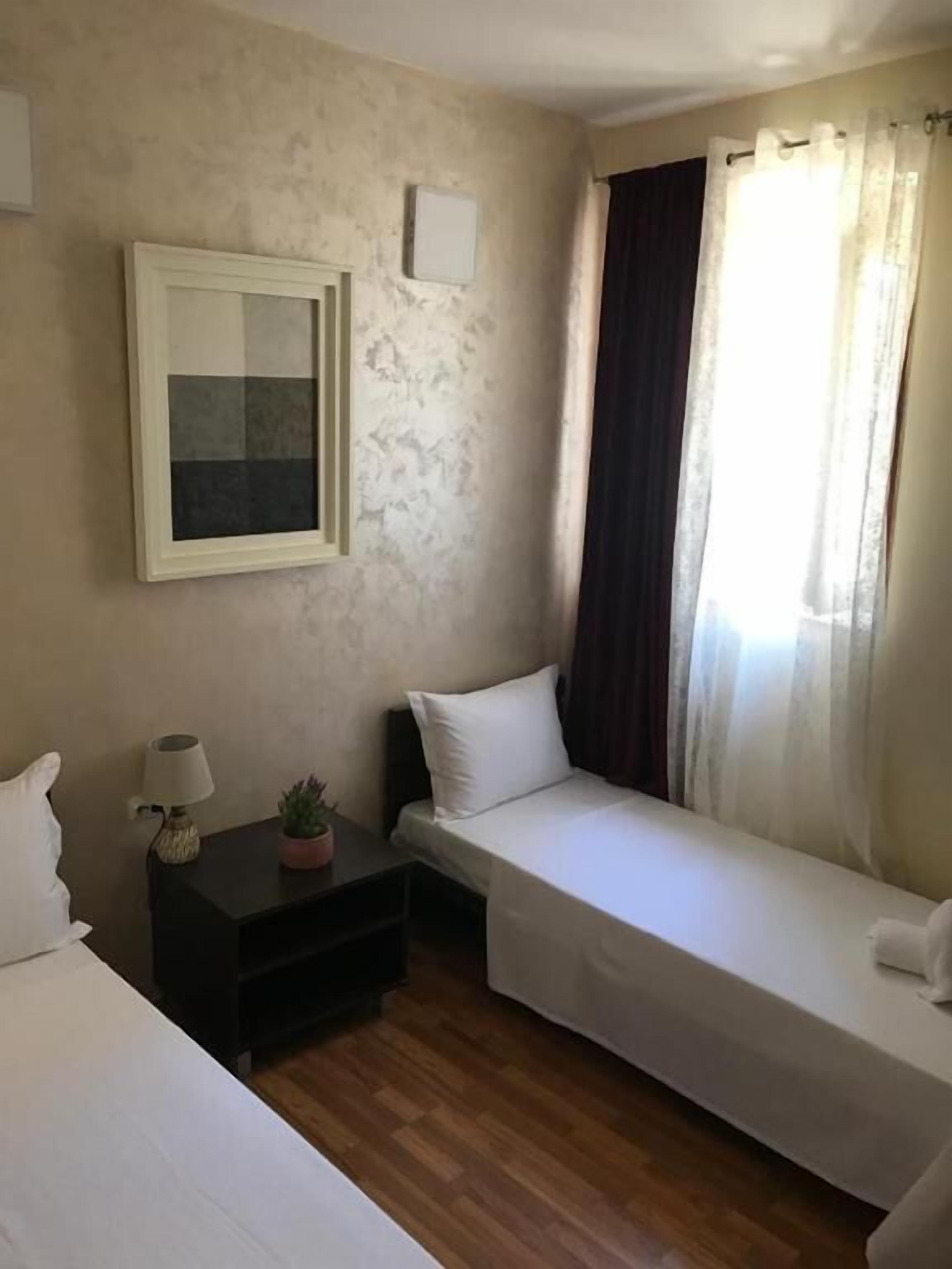 A small, simple room with two single beds, a nightstand with a lamp and a small plant, a framed picture on the wall, and a window with curtains.