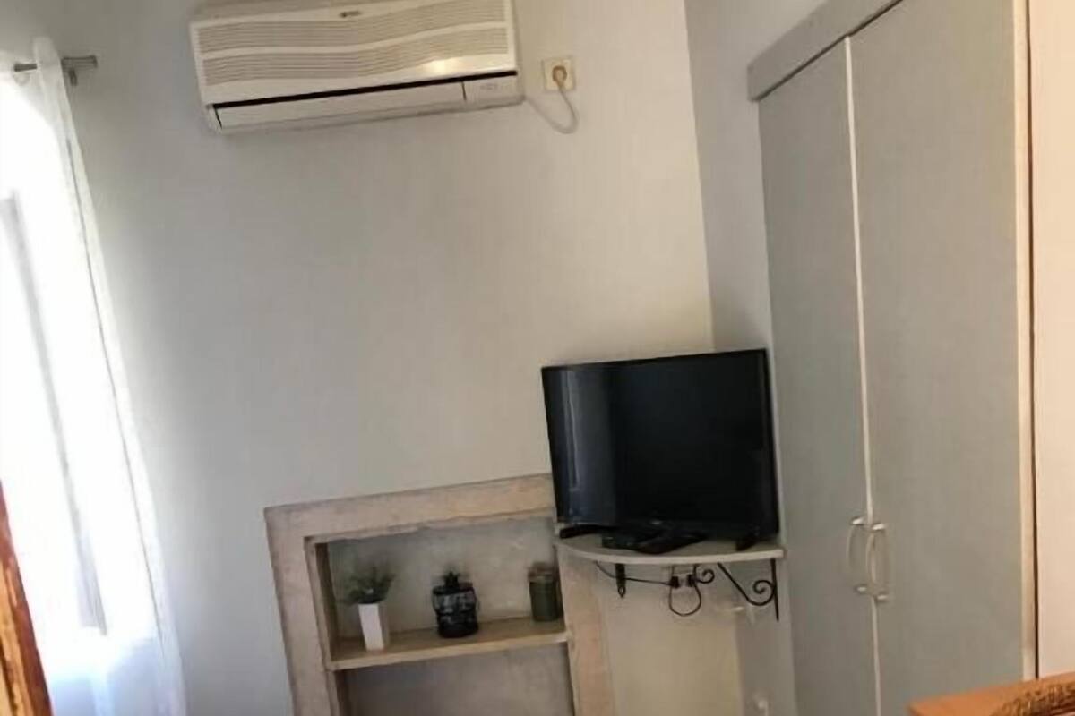 A small, tidy room with a bed, a TV on a shelf, a wardrobe, and a window with curtains.