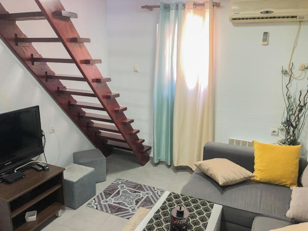 Ein Wohnzimmer mit einer Holztreppe, einem grauen Sofa mit gelben Kissen, einem Flachbildfernseher und einem gemusterten Teppich.