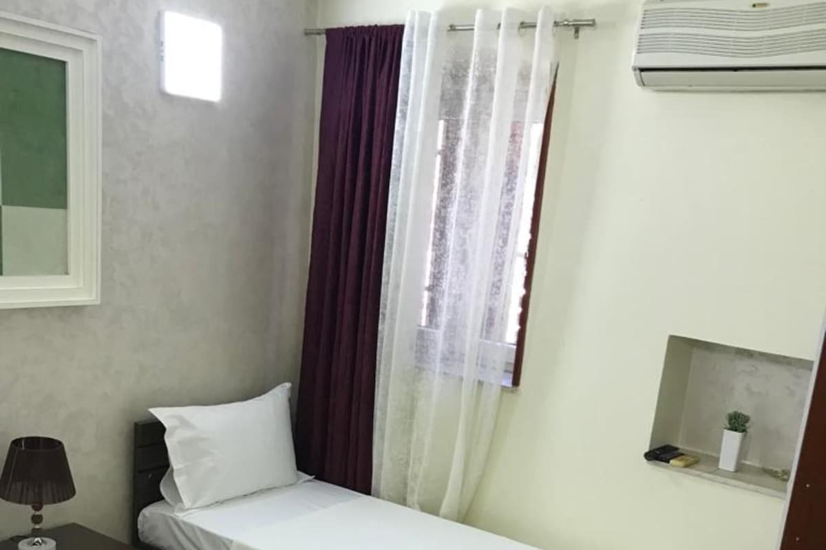 A small, clean room with a single bed, a nightstand, a desk, and a potted plant.