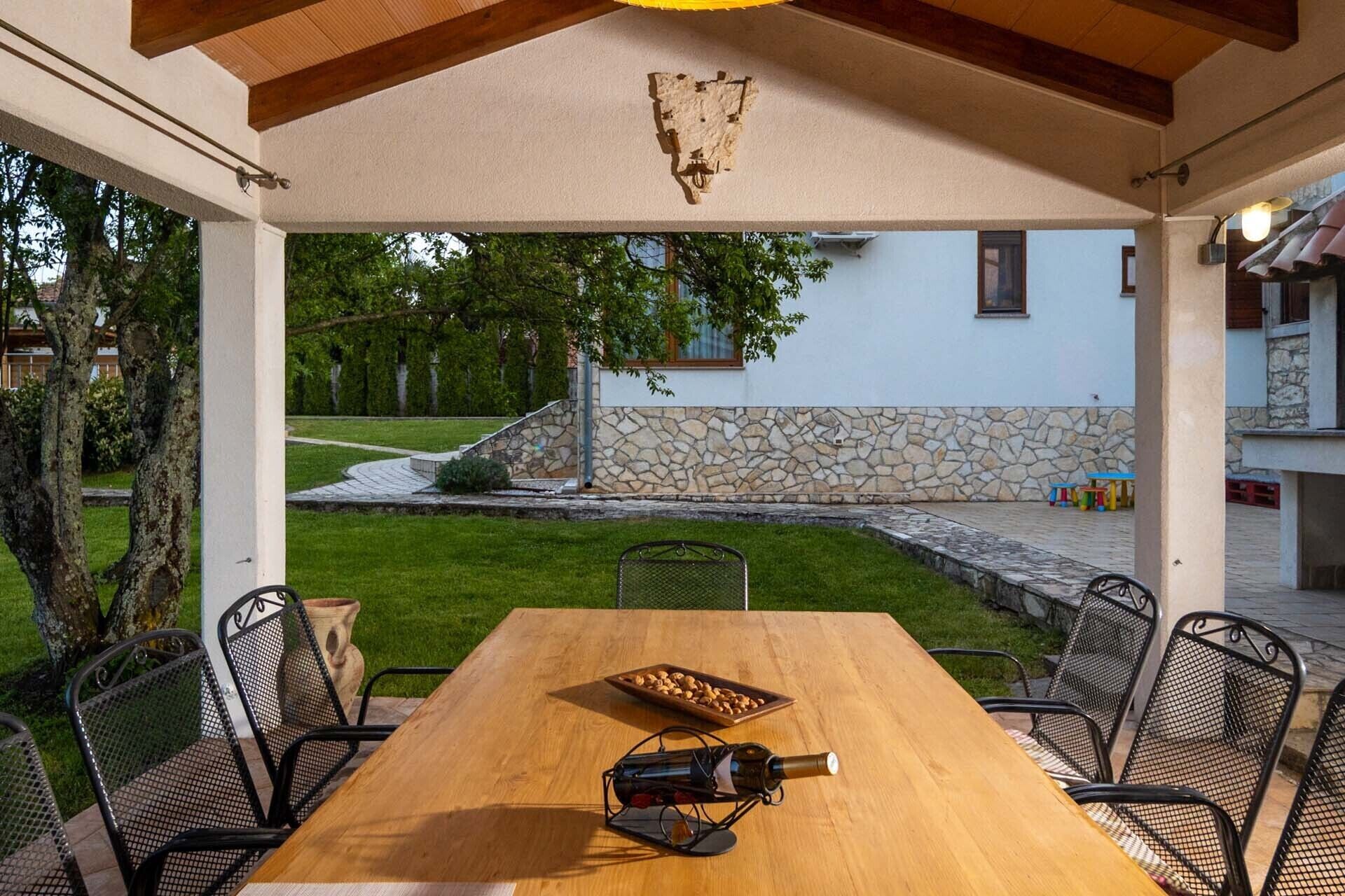 An outdoor patio with a wooden table, metal chairs, a wine bottle, and a stone wall in the background.