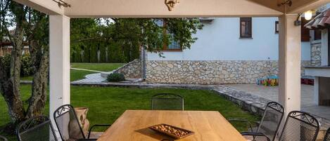 An outdoor patio with a wooden table, metal chairs, a wine bottle, and a stone wall in the background.
