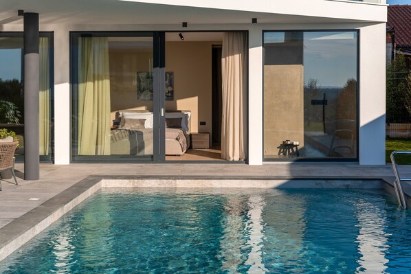 Une maison moderne dotée d’une piscine, de grandes portes vitrées donnant sur une chambre, et offrant une vue sur les environs.