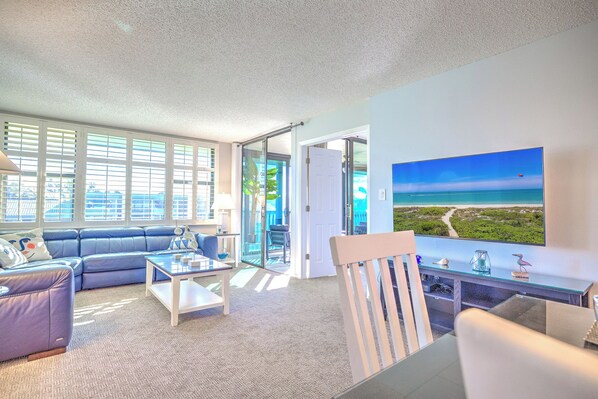 A modern living room with a large window, a blue sofa, a coffee table, and a painting of a beach view.