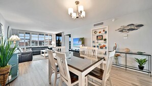 Een moderne woonkamer met een eethoek, grote ramen, uitzicht op zee en een decoratieve plank met diverse objecten.