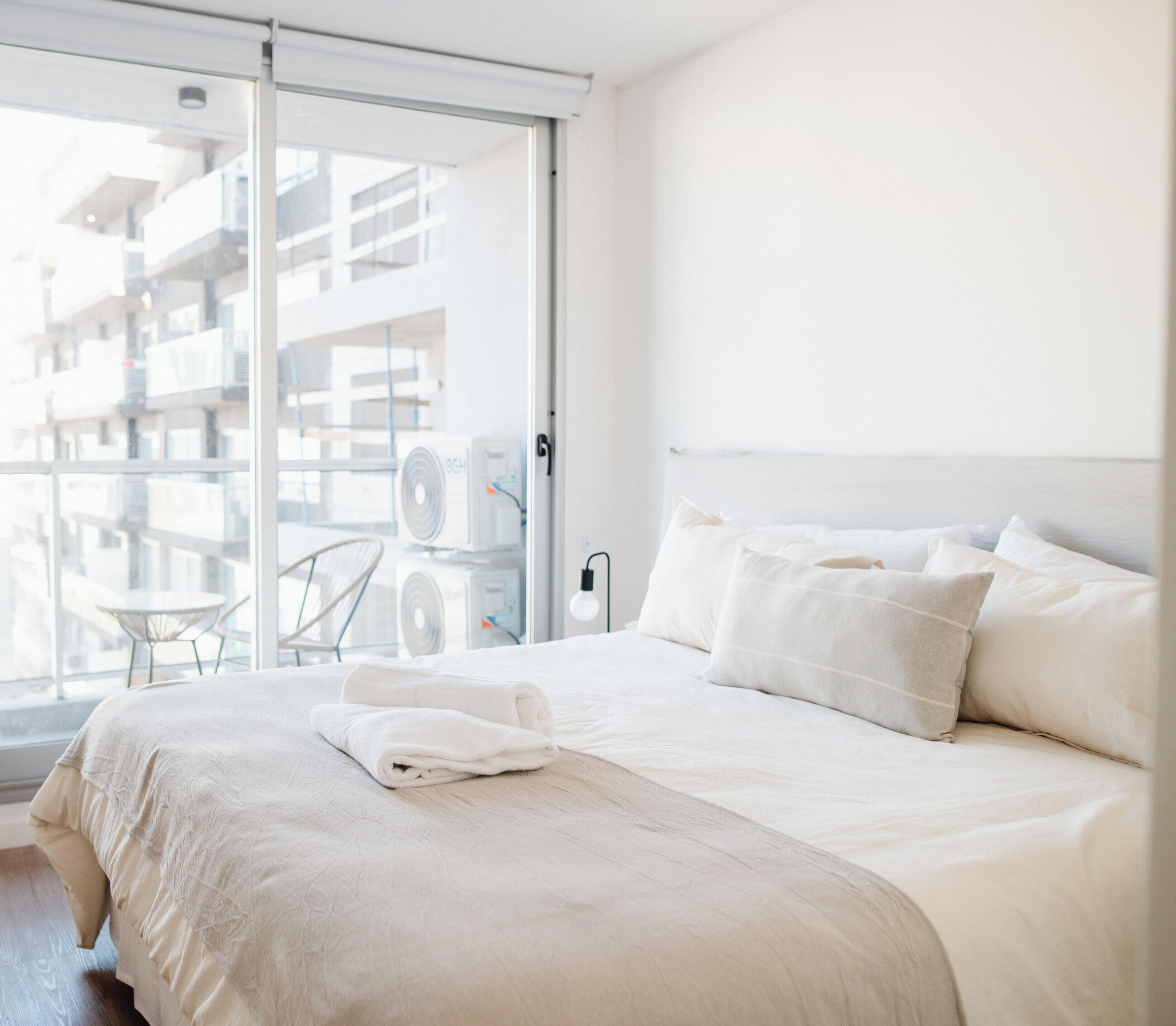 Ett hotellrum med en stor säng, utsikt över staden genom en skjutdörr och en balkong med utemöbler.