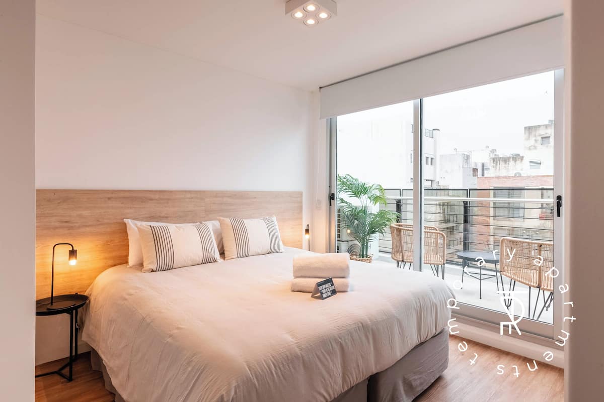 Un dormitorio moderno con una cama grande, una mesita de noche con lámpara, vistas de la ciudad a través de amplias ventanas y un balcón con sillas.