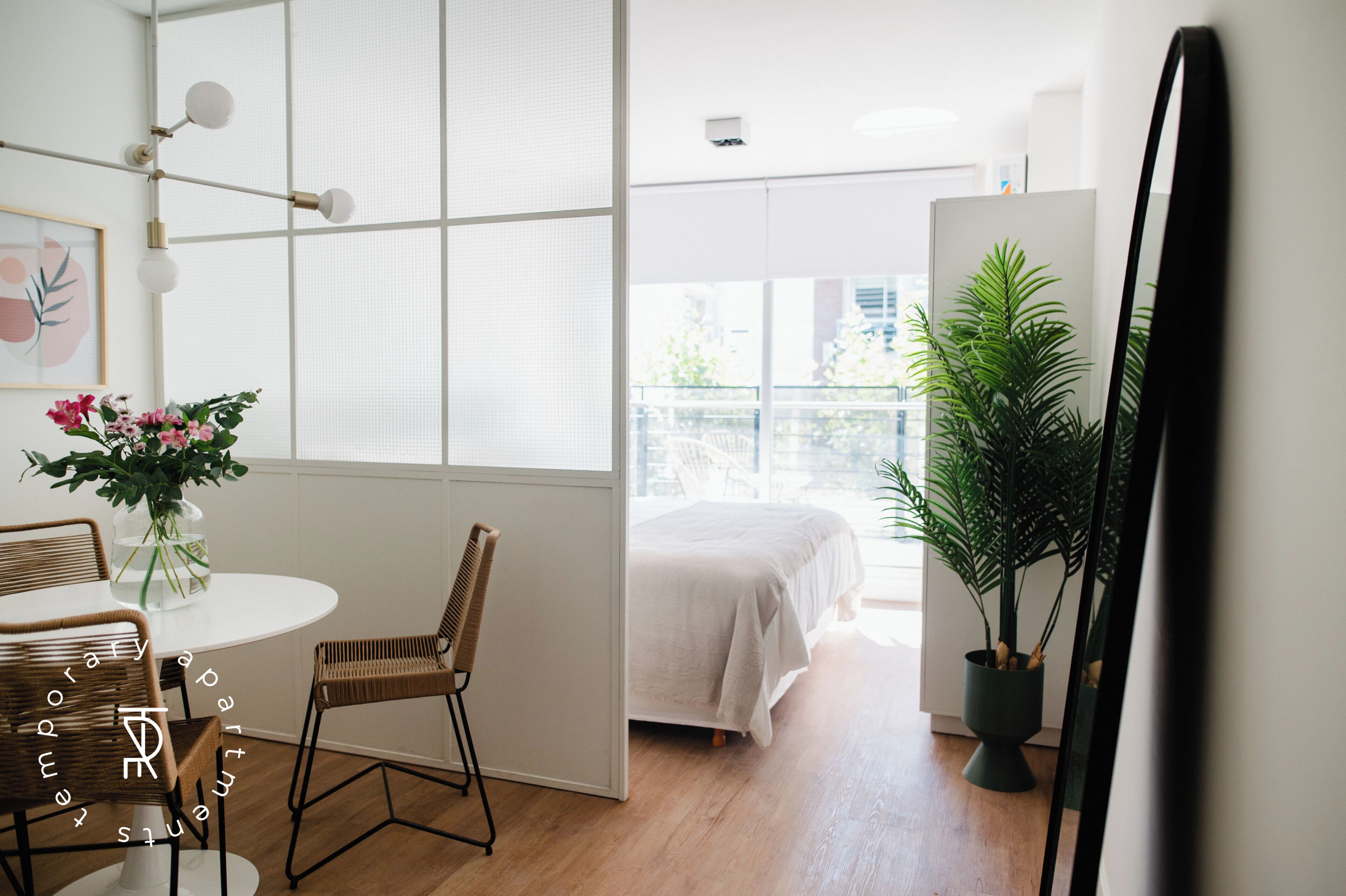 Ein Zimmer mit einem Bett, einem Esstisch mit Stühlen, einer Topfpflanze und einem großen Spiegel.