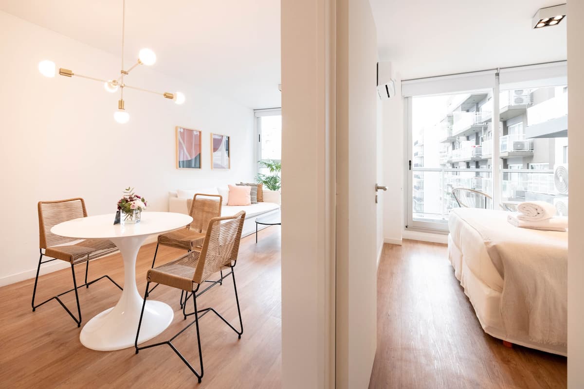Habitación de hotel moderna con una mesa redonda para comer, sillas de mimbre, cama con edredón blanco y un balcón con vista a la ciudad.
