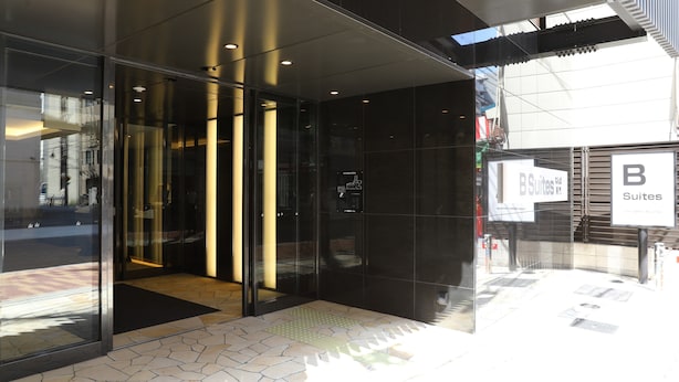 Entrada de un hotel con puertas de vidrio, un letrero que dice 'B Suites' y un piso de baldosas.