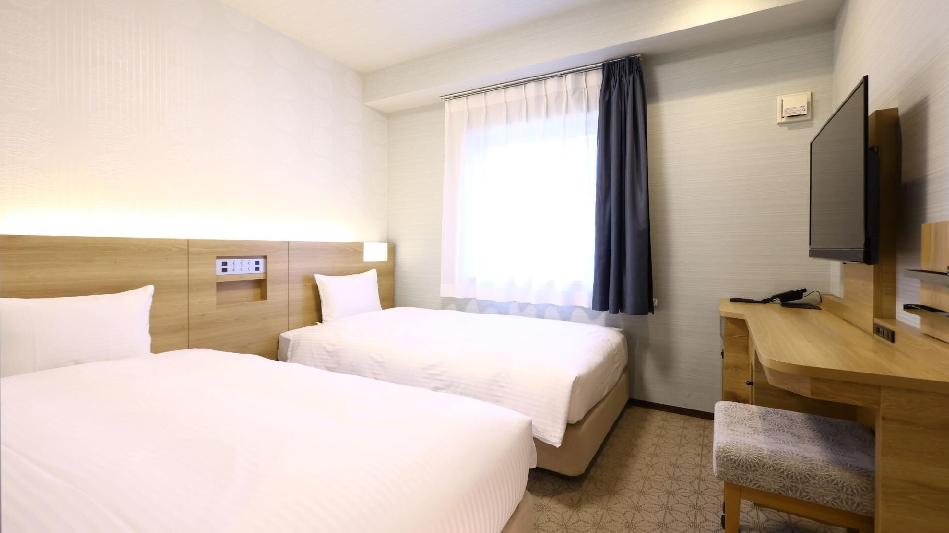 A hotel room with two beds, a desk, a TV, and a window with curtains.