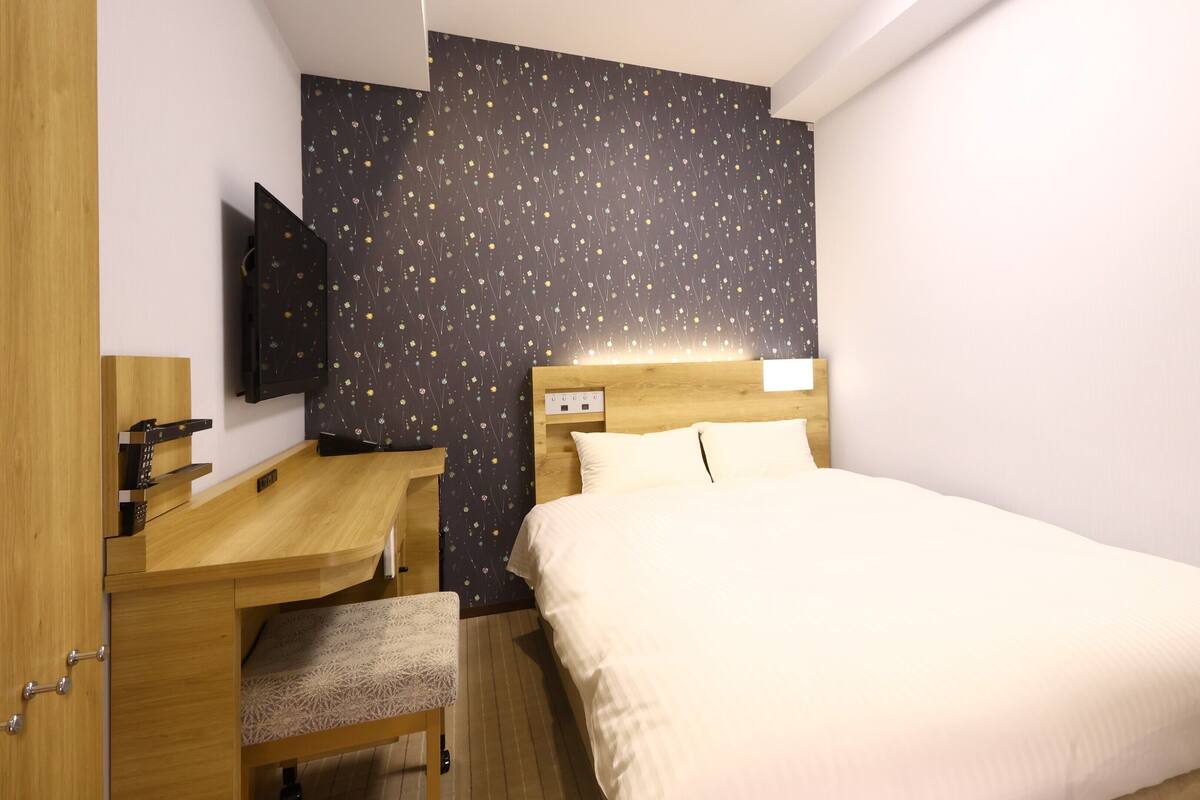 Habitación de hotel con cama, escritorio, silla, televisor y una pared con estampado floral.