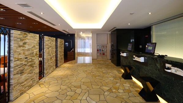 A hotel reception area with a stone-tiled floor, a reception desk with a computer, and a glass partition.
