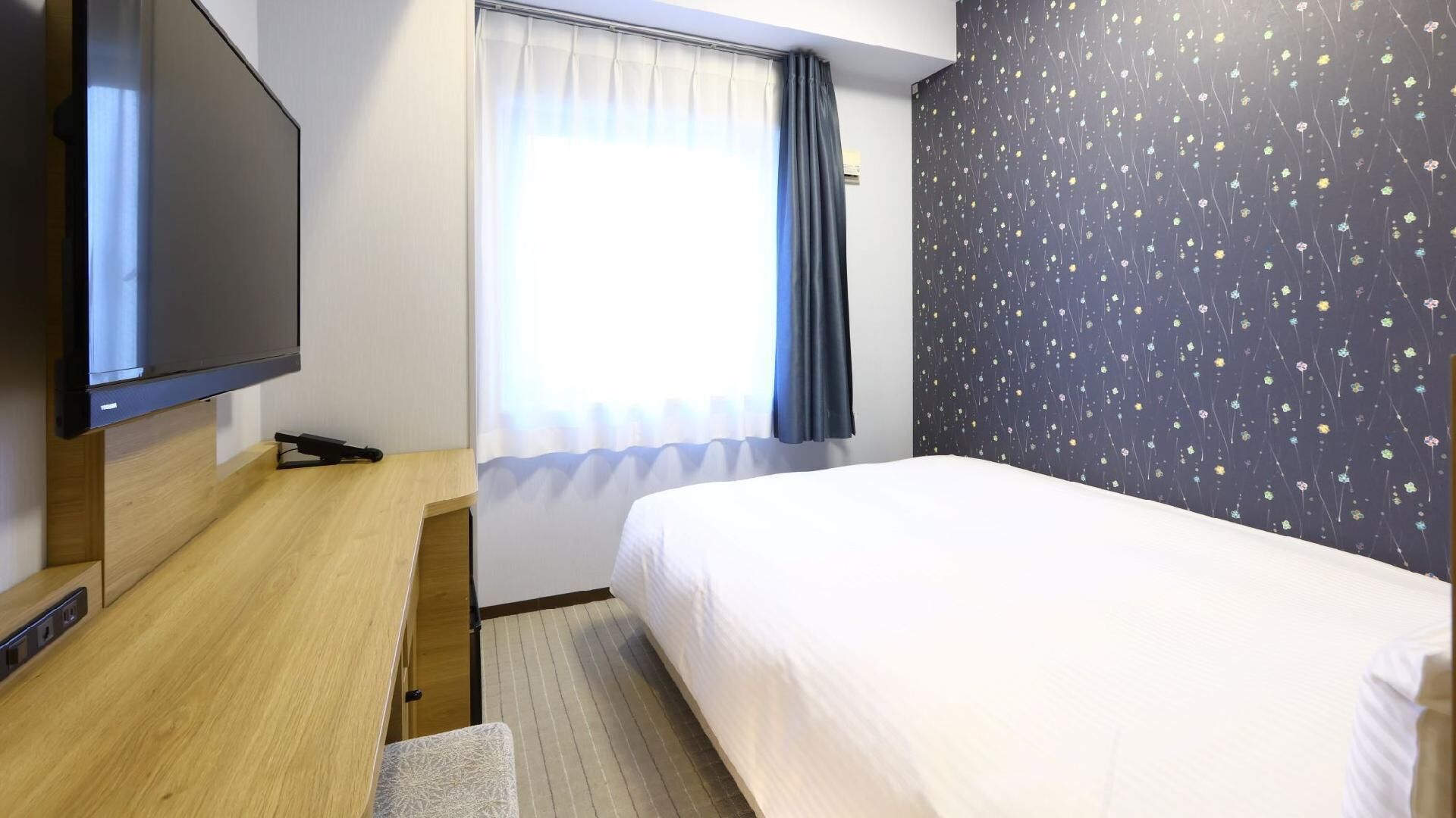 A hotel room with a bed, a TV, a desk, and a window with curtains.