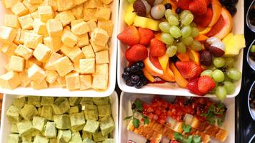 Four trays of food: cubes of yellow cheese, a mix of fruits and grapes, cubes of green cheese, and a dish with bread, tomatoes, and greens.