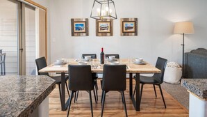 A dining area with a wooden table, black chairs, a hanging light fixture, and framed pictures on the wall.