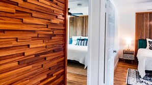 A hotel room with a wooden wall, a bed with white bedding, a nightstand with a lamp, and a view into another room. - 400 South - Unit E (Cocoa Beach)