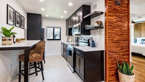 A modern kitchen with dark cabinetry, a wooden accent wall, and a dining area with wicker chairs. - 400 South - Unit E (Cocoa Beach)