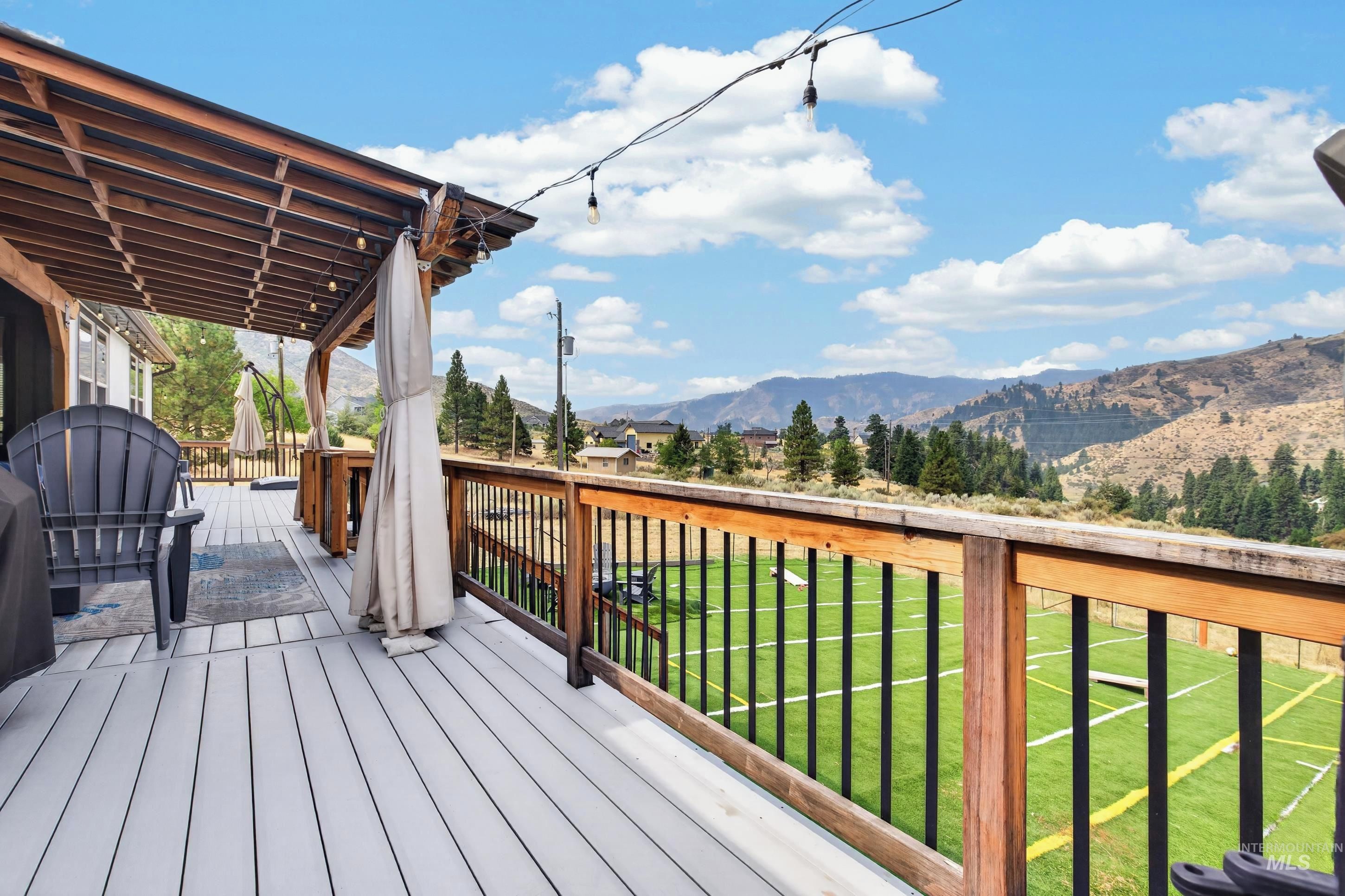Una terraza de madera con mobiliario exterior, un pérgola y vistas a un campo verde y montañas.