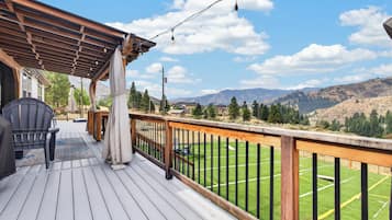 Una terraza de madera con mobiliario exterior, un pérgola y vistas a un campo verde y montañas.