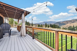 A wooden deck with outdoor furniture, a pergola, and a view of a green field and mountains.