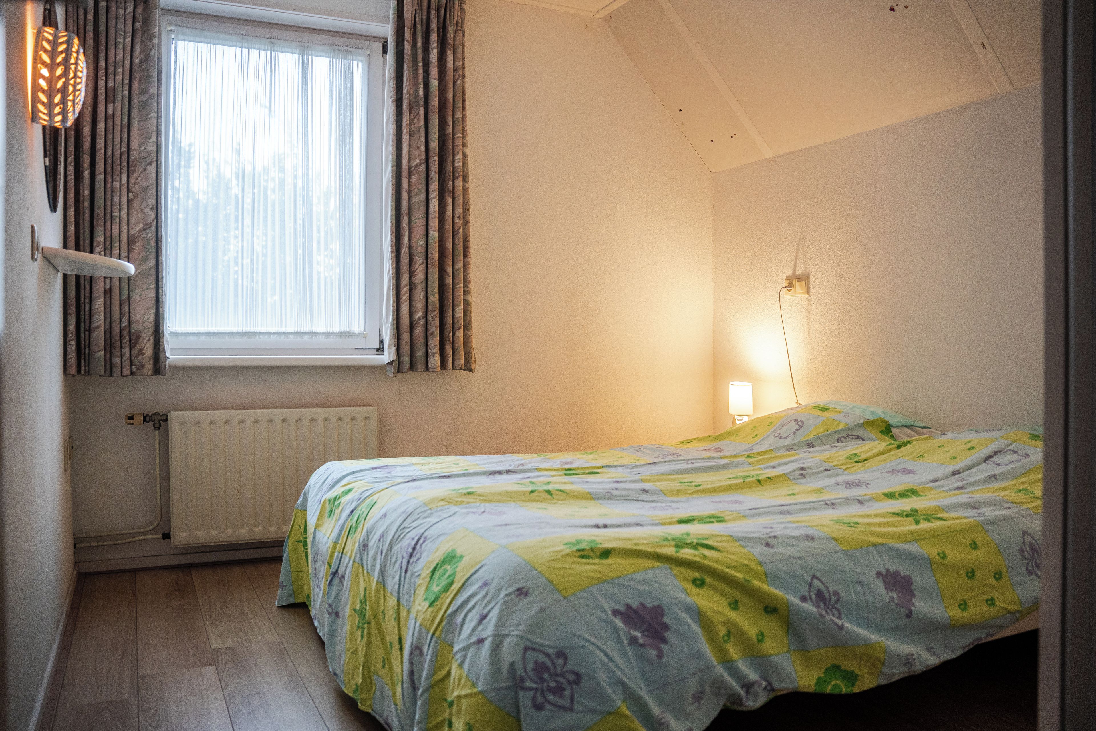 A bedroom with a bed, a radiator, a window with curtains, and a wall-mounted light fixture.