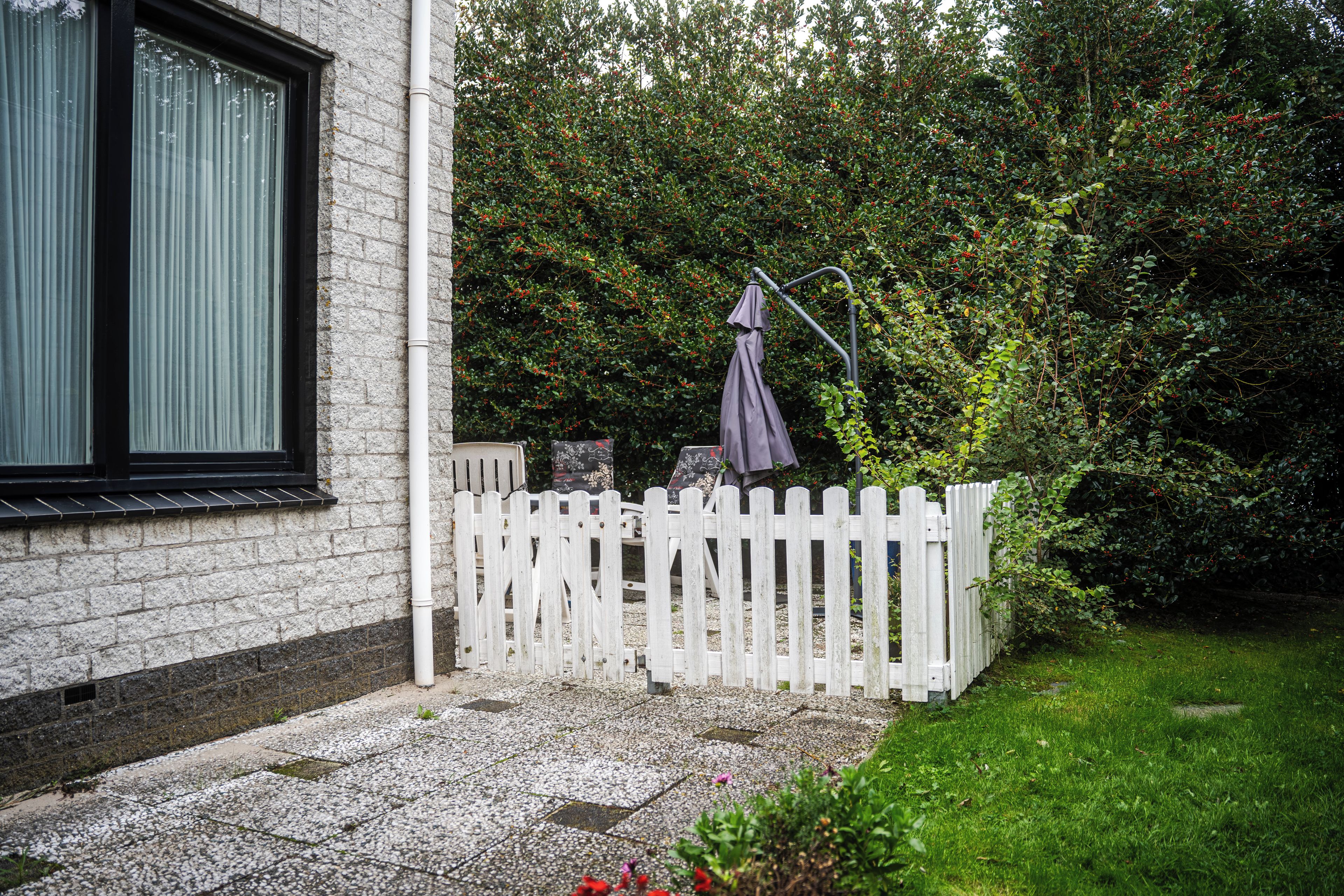 A white picket fence, a window with blinds, and a garden with a metal umbrella stand.