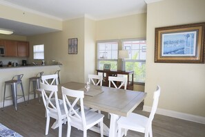 A dining area with a table and white chairs, a kitchen in the background, and a framed picture on the wall.