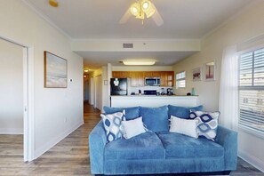 Sala de estar con un sofá azul y cojines estampados, una cocina al fondo y un ventilador de techo.