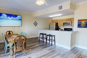 A modern kitchen with wooden cabinets, a stainless steel refrigerator, and a microwave. There is a dining area with a wooden table and wicker chairs.