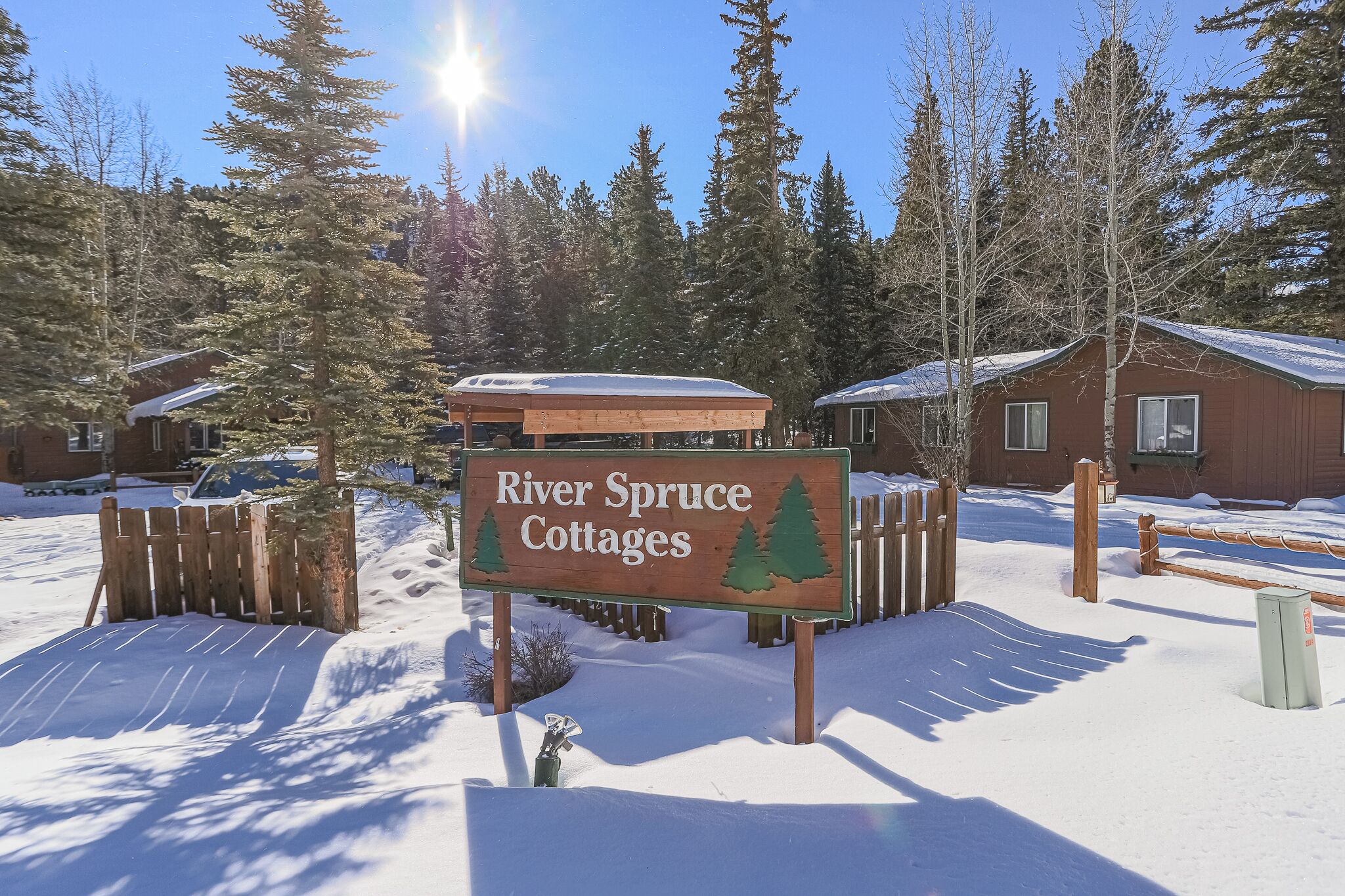 A sign for River Spruce Cottages with a wooden fence and snow-covered ground.