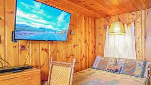 A cabin interior with a television mounted on the wall, a bed with a forest-themed bedspread, and a wicker chair. - Original RMNP Riverfront Cabin on the Thompson River (Estes Park)