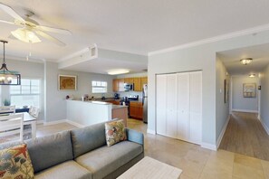 A modern living room with a gray sofa, a dining area with a white table, and a kitchen with wooden cabinets and stainless steel appliances.