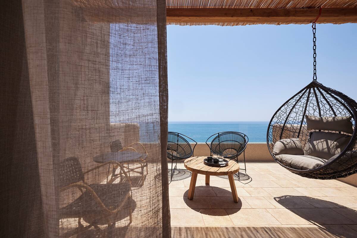 A patio with a hanging chair, a round table, and chairs facing a view of the sea.