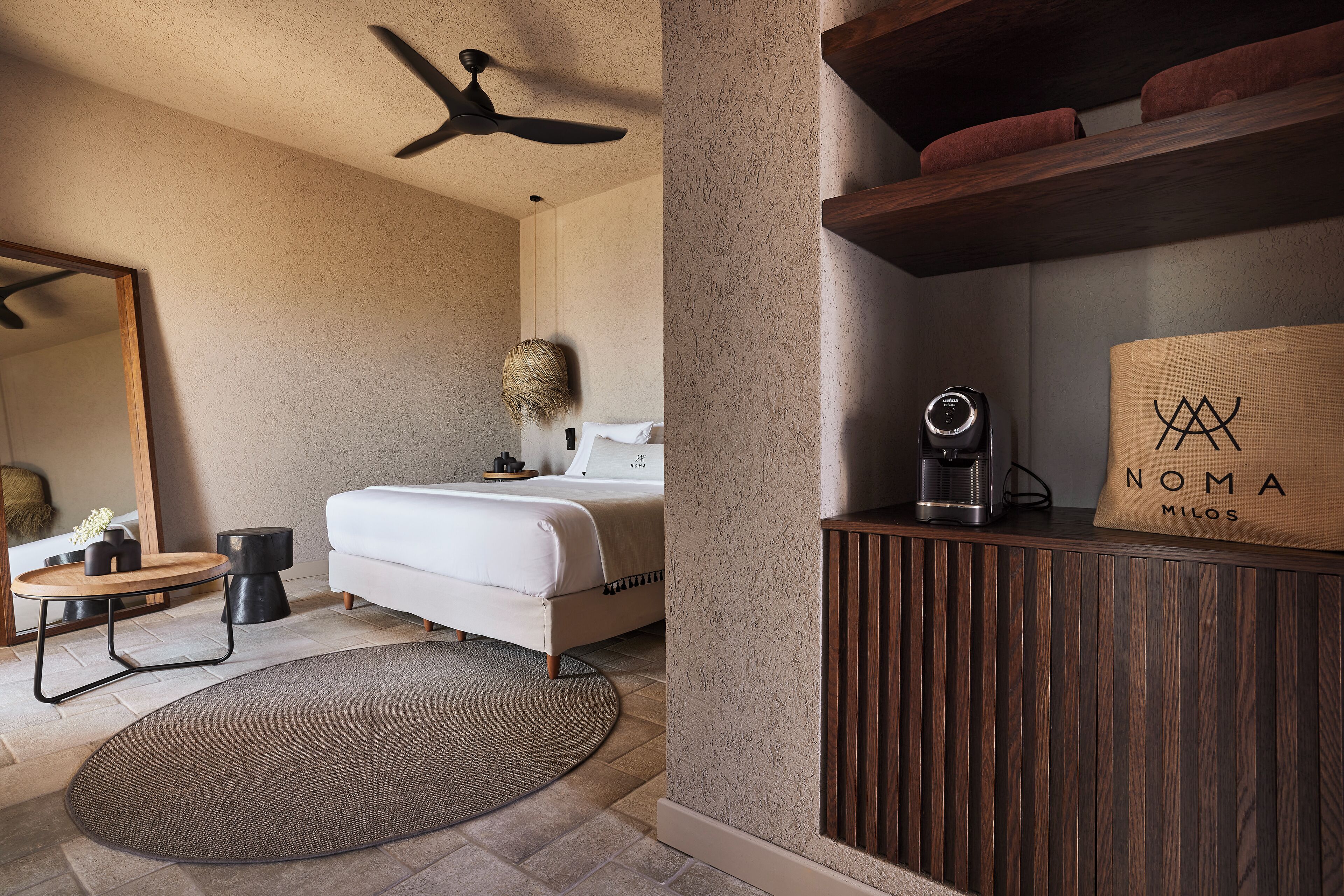 Ein modernes Hotelzimmer mit Bett, runder Teppich, Holz-Nachttisch, Spiegel und Deckenventilator.