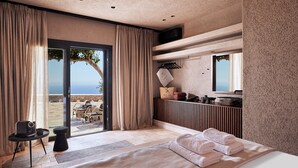 A modern bedroom with a large bed, a seating area with a table, a view of the outdoors, and a bathroom with a mirror and towel rack. - Noma Milos - Delmar Collection (Milos)