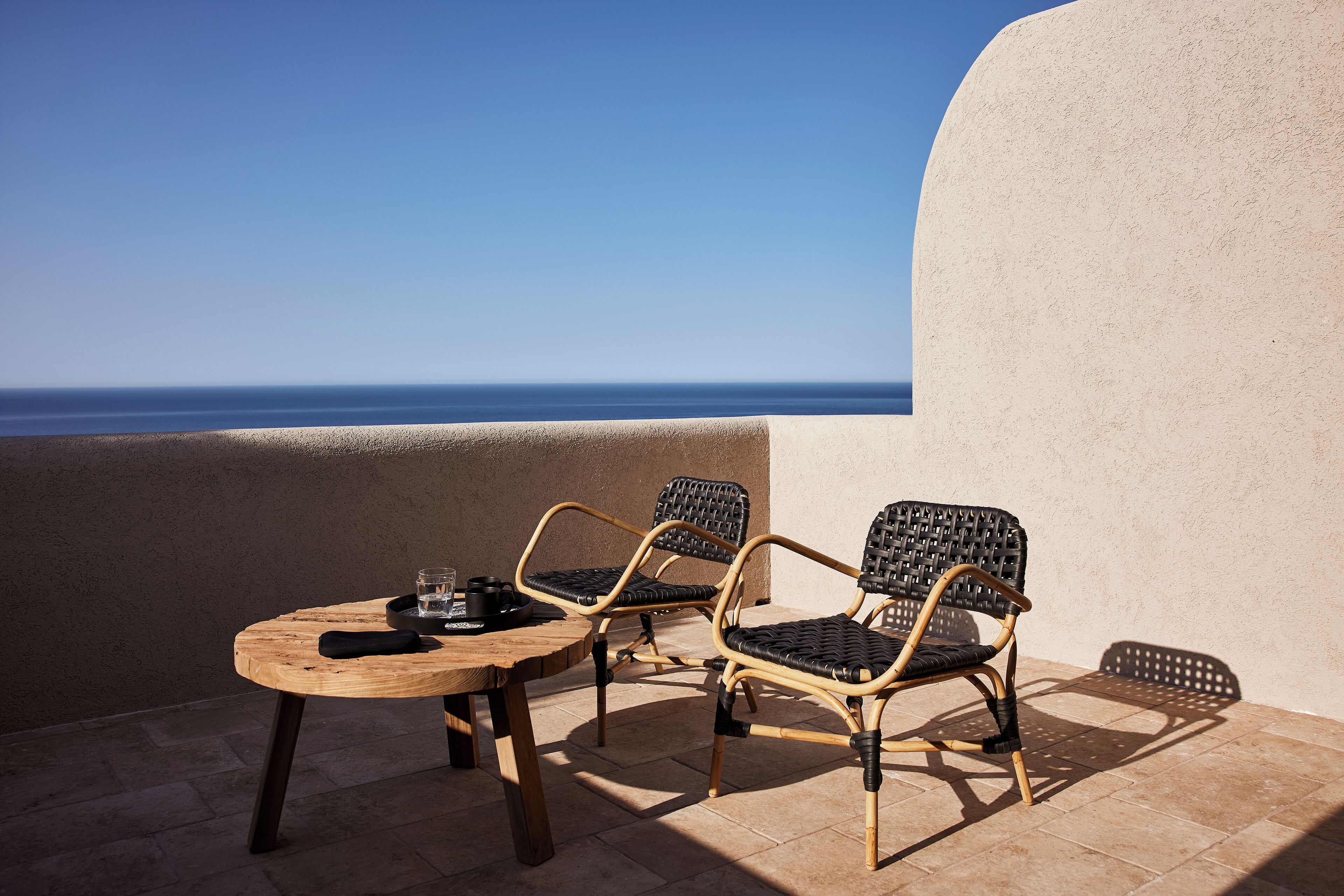 Eine Terrasse mit einem Holztisch und zwei Korbstühlen, mit Blick auf einen klaren, blauen Himmel und das Meer.