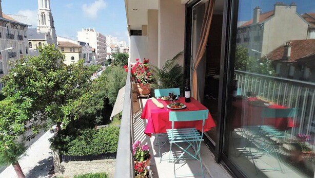 A balcony with a red table and chairs, a potted plant, and a view of buildings and a church. - T3 with parking 65 m2, 5 min walk from the beach. (Biarritz)
