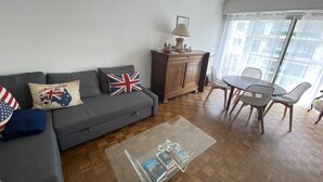 A living room with a grey sofa, a glass coffee table with books, a wooden cabinet, and a dining area with chairs and a table. - T3 with parking 65 m2, 5 min walk from the beach. (Biarritz)