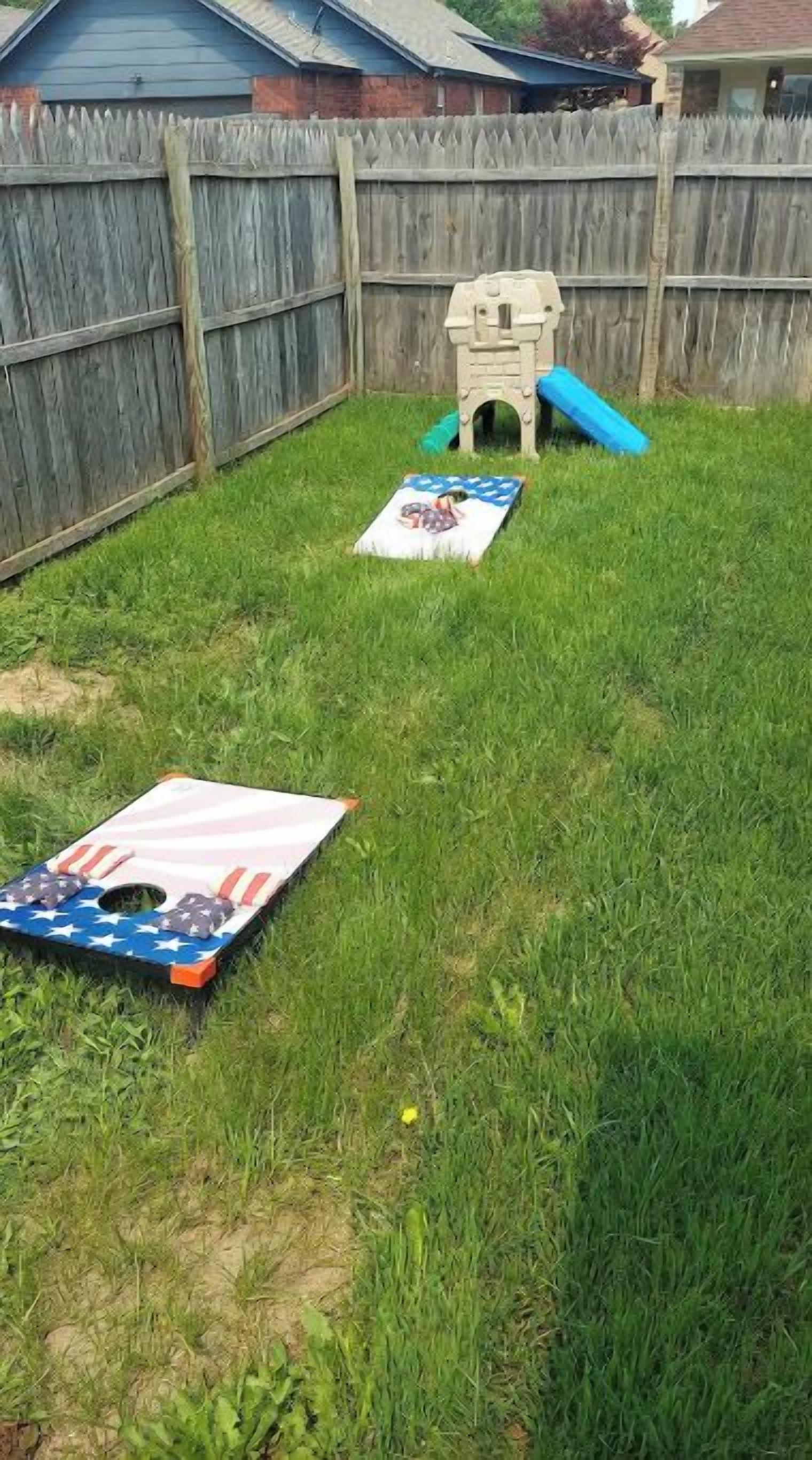 A grassy backyard with a wooden fence, a small play structure, and a mat with an American flag design.