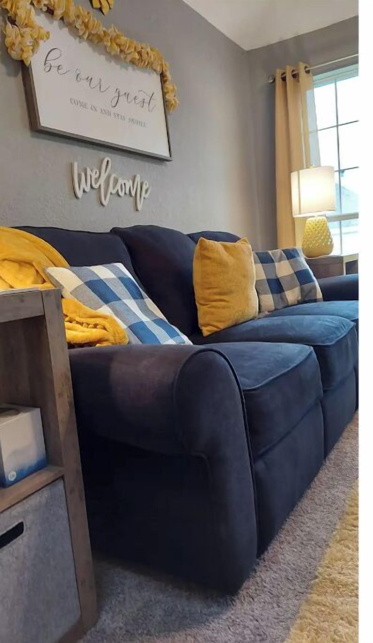 A living room with a dark blue sofa, yellow and blue pillows, a wooden side table, a framed sign on the wall, and a lamp on a side table.