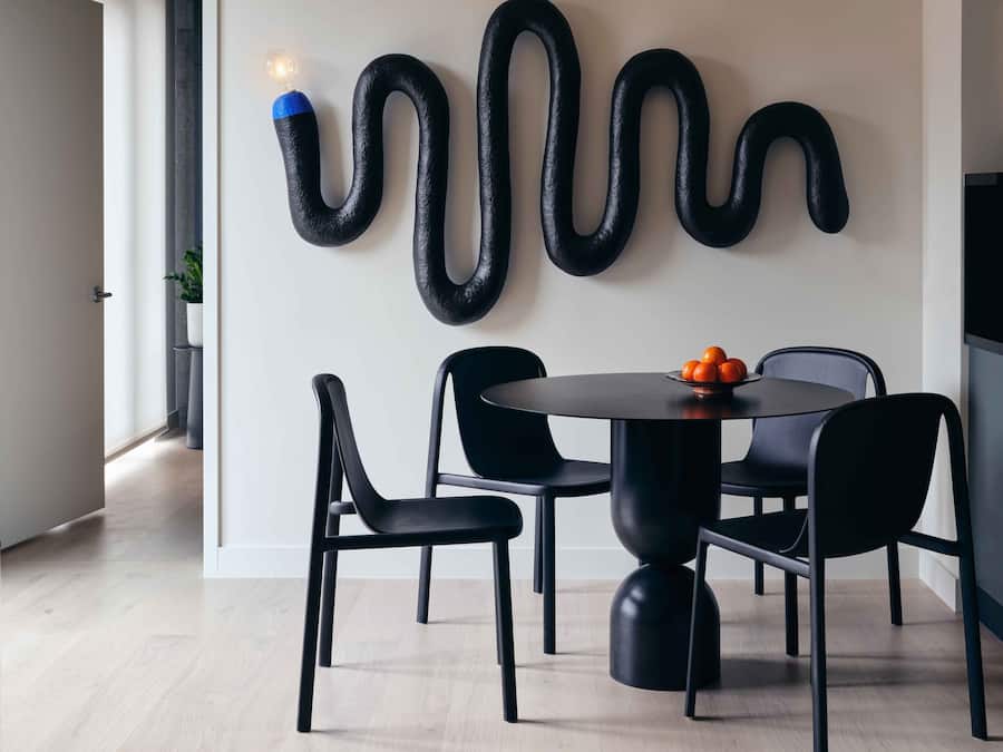 Een moderne eetruimte met een ronde tafel, zwarte stoelen en een abstracte sculptuur aan de muur.