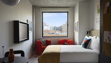 A hotel room with a large window, a bed with multiple pillows, a small table with a bottle, and a chair.